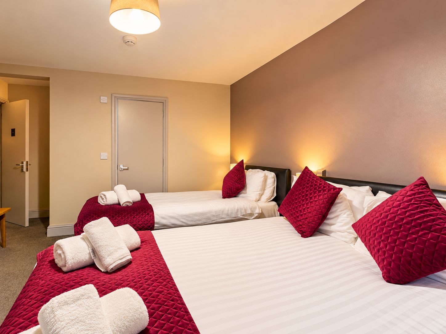 A hotel room with two beds, each with white linens and red pillows, and a red quilted bedspread. There are rolled towels on the beds and a door in the background.