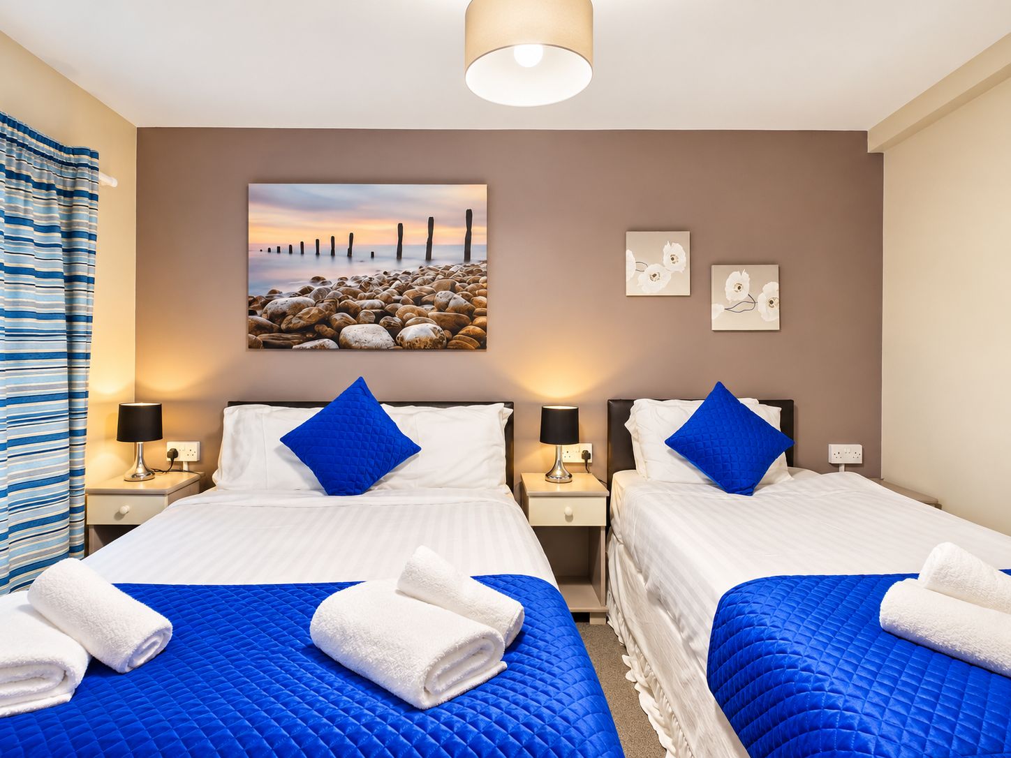 Habitación de hotel con dos camas, cada una con cubrecama azul acolchado y ropa de cama blanca. Hay dos lámparas de pared a cada lado de las camas, y un cuadro grande con una escena de playa sobre ellas.