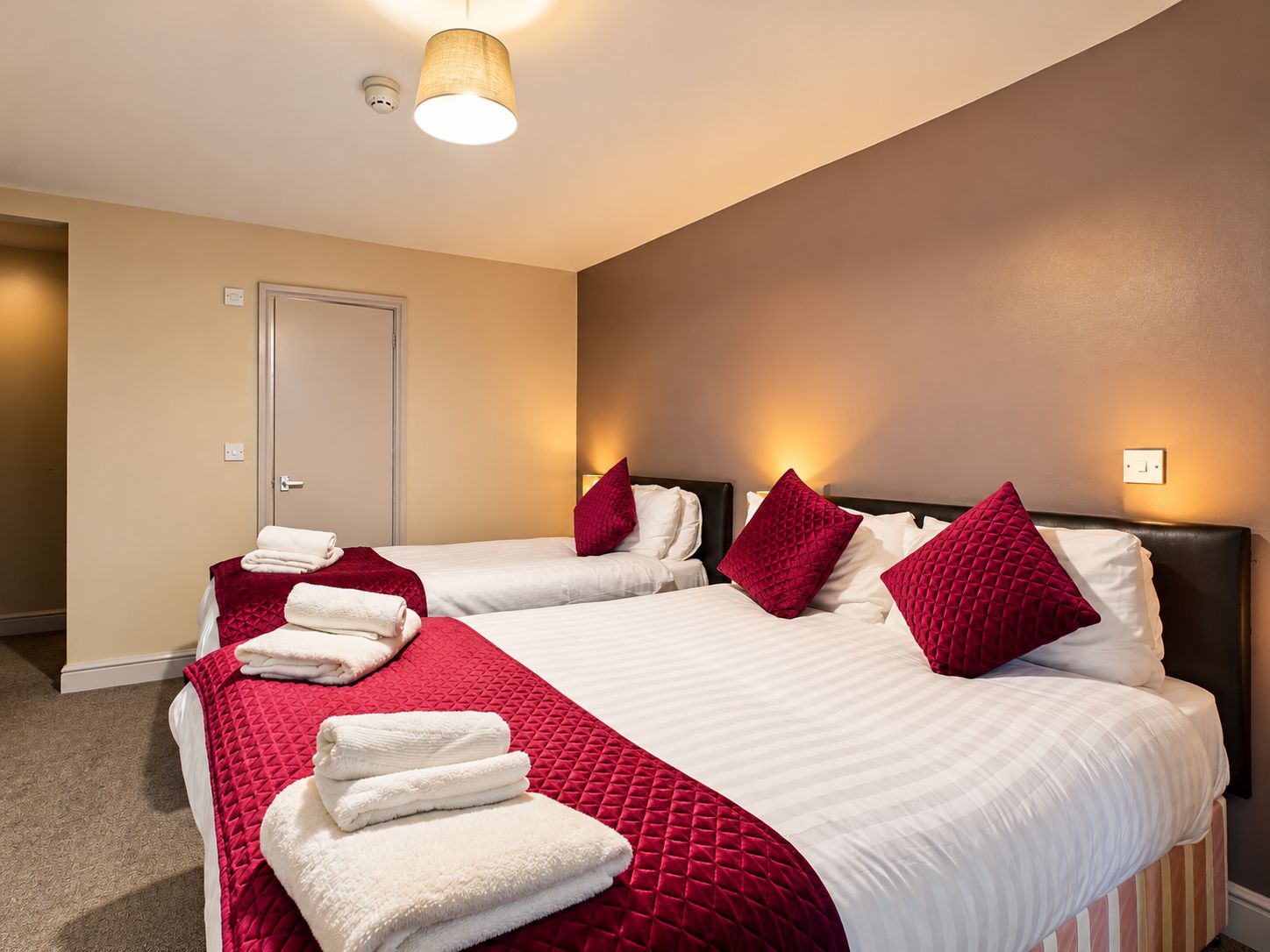 Habitación de hotel con dos camas, cada una con sábanas blancas y almohadas rojas, y una colcha acolchada roja. Hay toallas blancas dobladas sobre las camas, y la habitación tiene paredes color beige y un accesorio de iluminación en el techo.