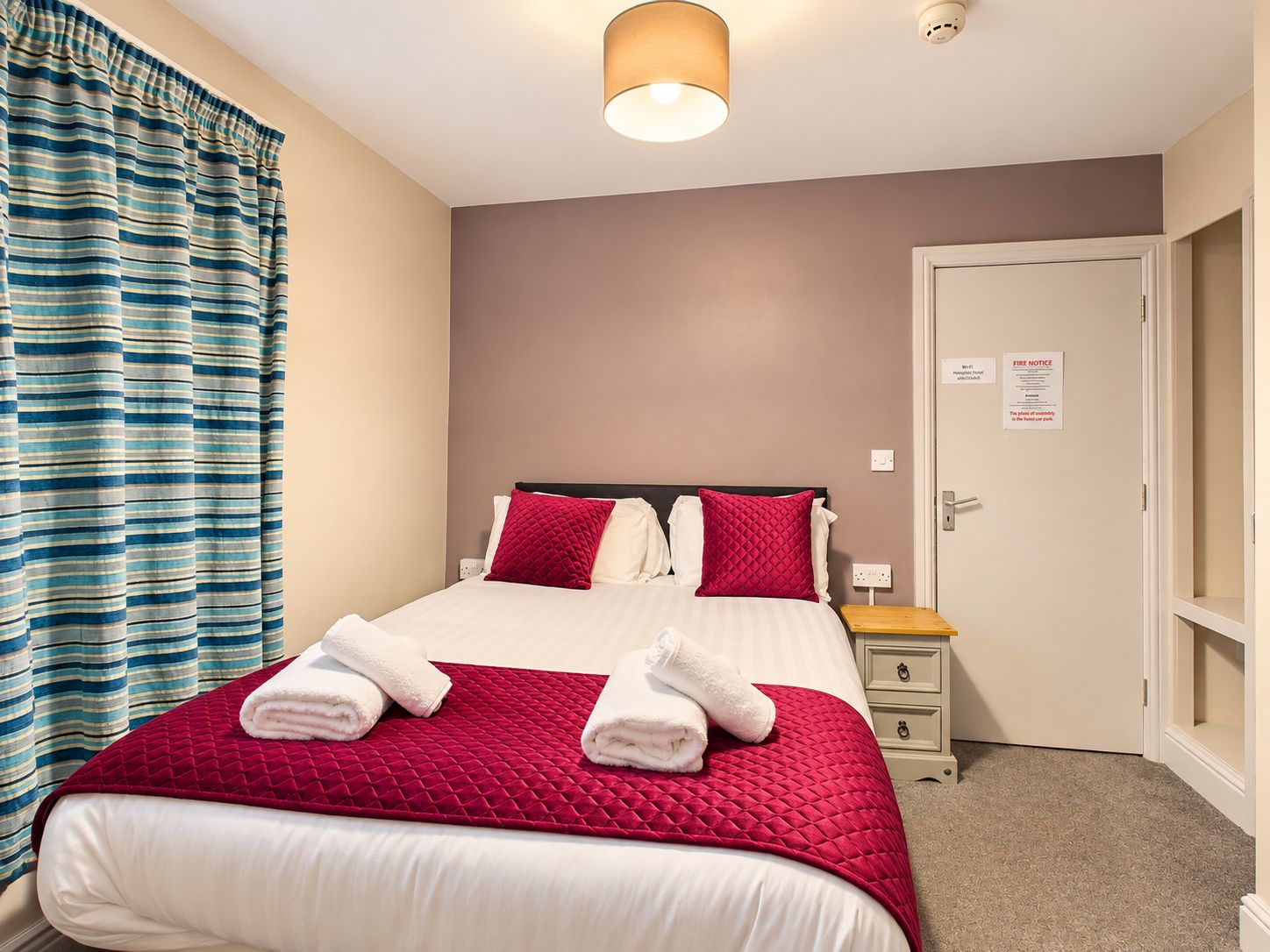 Una habitación de hotel con cama, mesita de noche, puerta y ventana con cortinas.