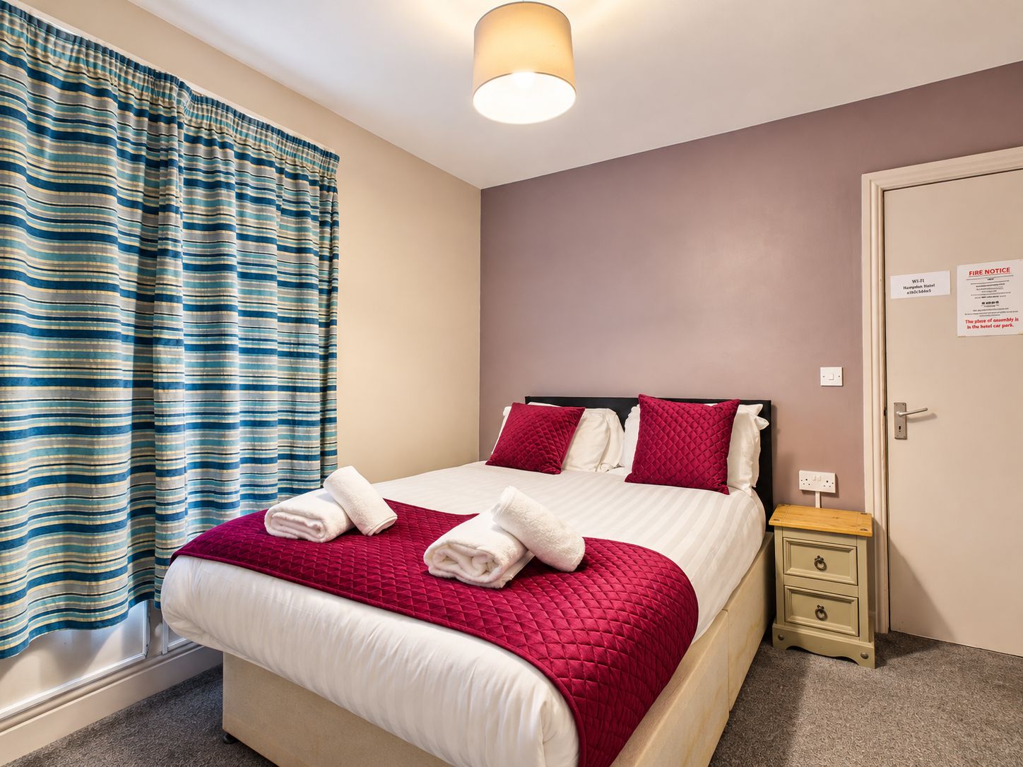A hotel room with a bed, a nightstand, a lamp, a window with curtains, and a door with a fire notice.