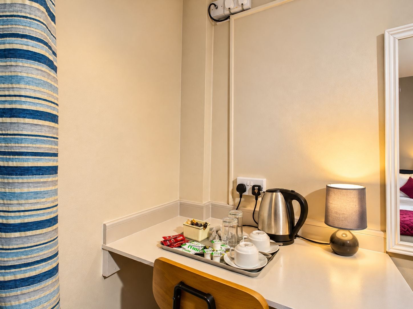 A hotel room with a kitchenette, a kettle, a tray with food items, and a lamp.