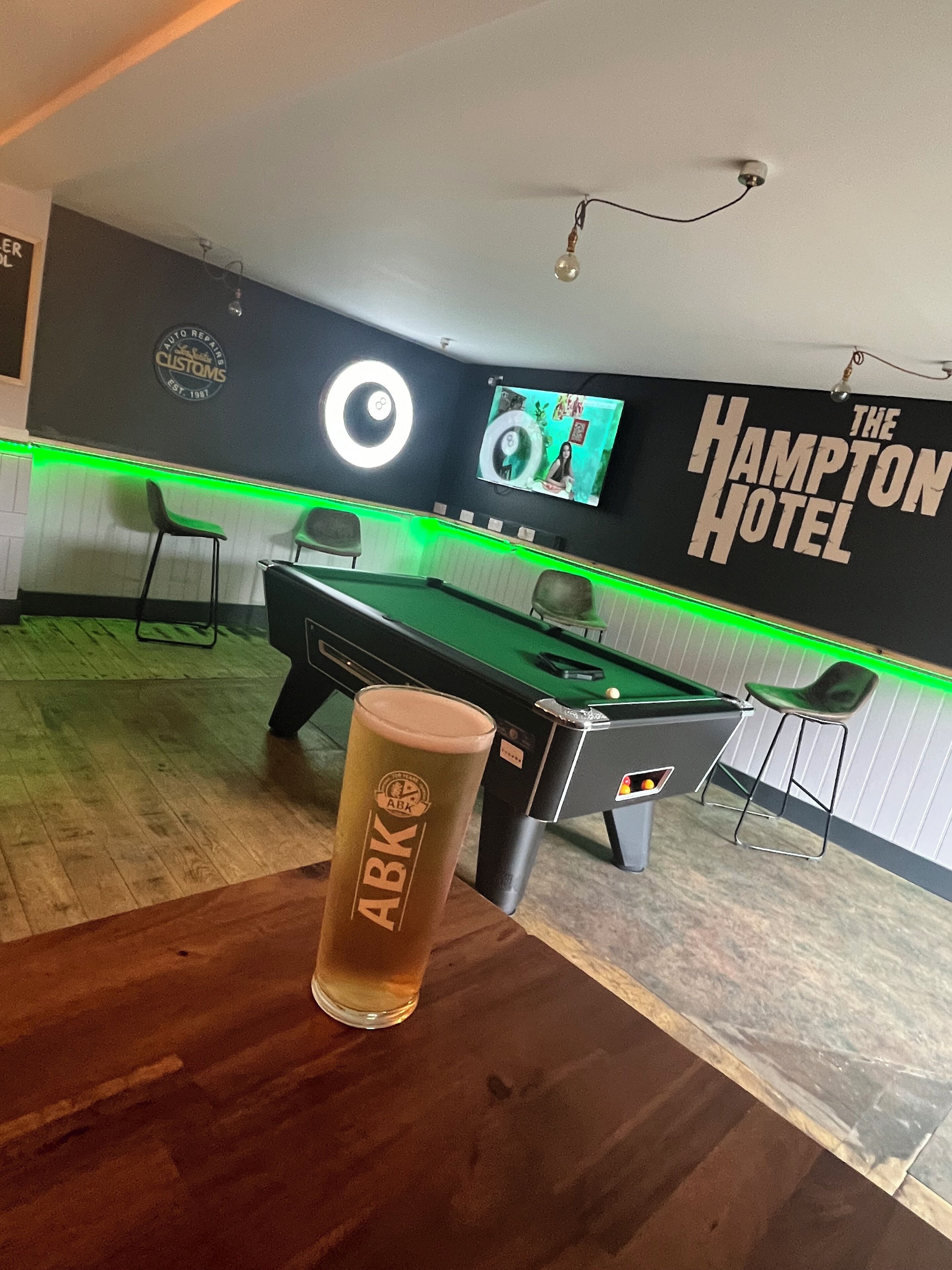 Una mesa de billar con taco, una cerveza en vaso y una pared con el letrero 'The Hampton Hotel'.