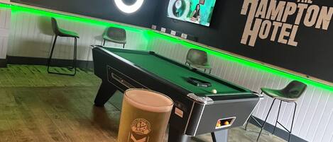 Una mesa de billar con taco, una cerveza en vaso y una pared con el letrero 'The Hampton Hotel'.