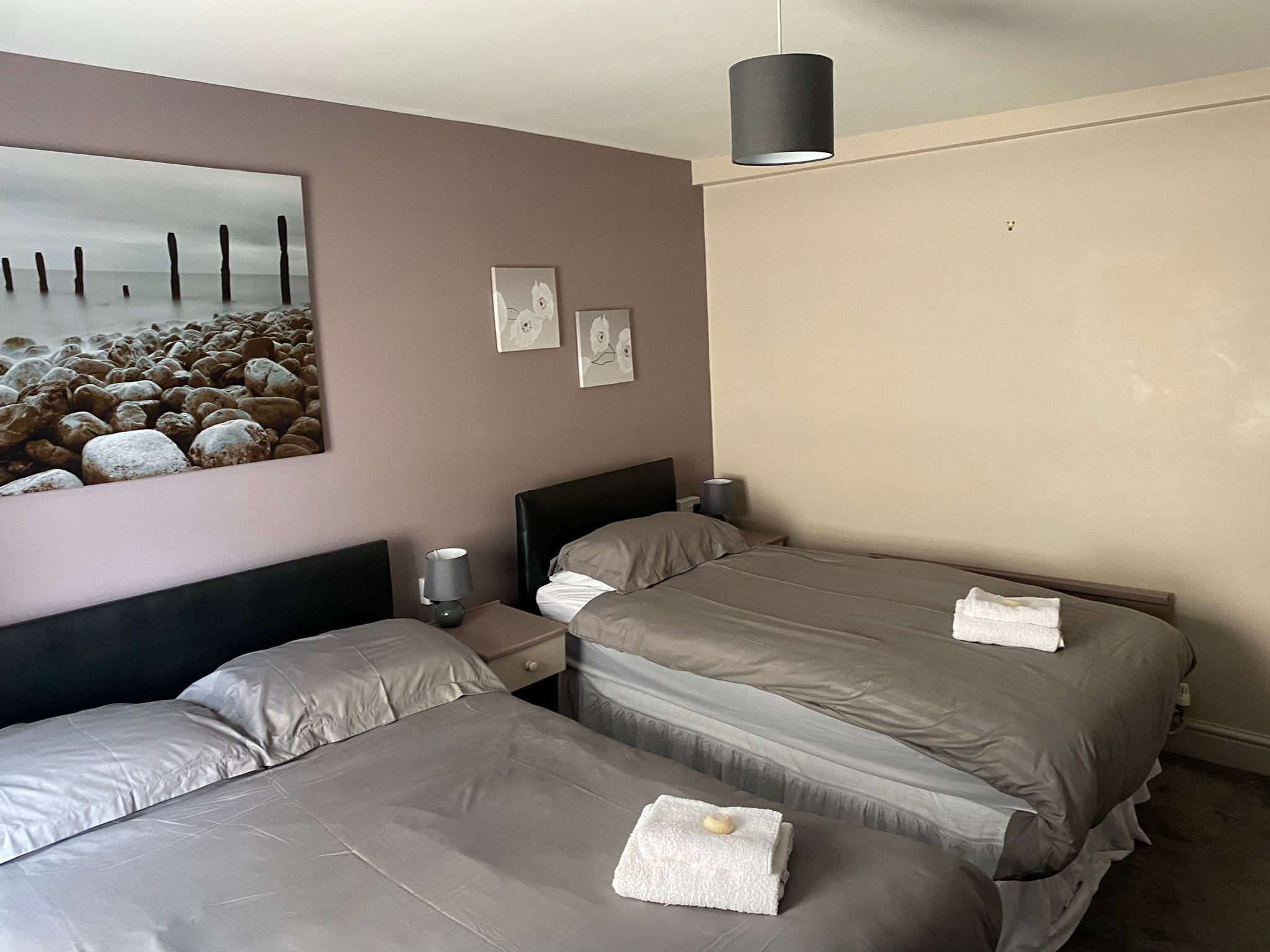 Habitación de hotel con dos camas, una mesita de noche con lámpara, un cuadro con escena de playa y una lámpara de techo.