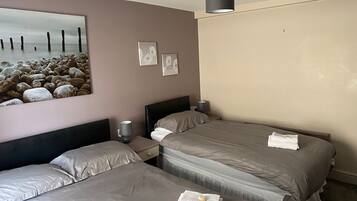 A hotel room with two beds, a nightstand with a lamp, a painting of a beach scene, and a ceiling light.