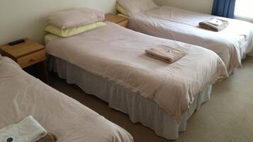 A hotel room with three single beds, each with a pillow and a folded towel. There is a wooden nightstand with a remote control on it.