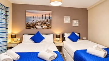 A hotel room with two beds, each with a blue quilted bedspread and white linens, a painting of a sunset over rocks on the wall, and a single light fixture above.