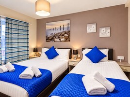 Habitación de hotel con dos camas, ropa de cama azul, toallas blancas, un cuadro de una playa y una ventana con cortinas.