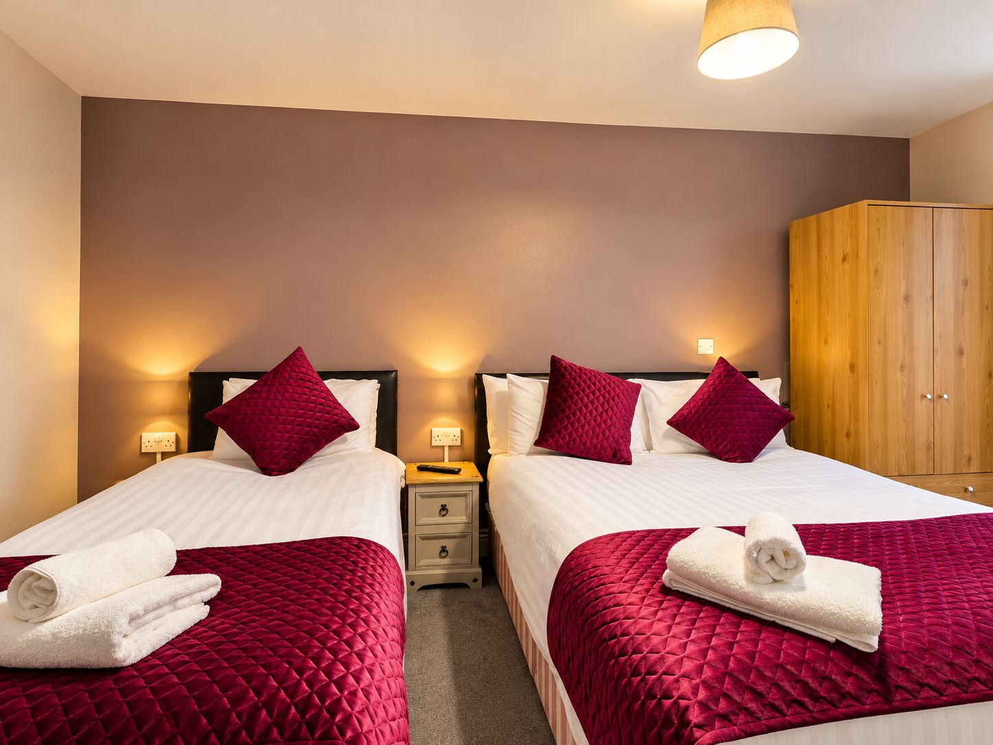 Habitación de hotel con dos camas, cada una con cubrecama rojo acolchado y almohadas blancas, un armario de madera y lámparas de noche.