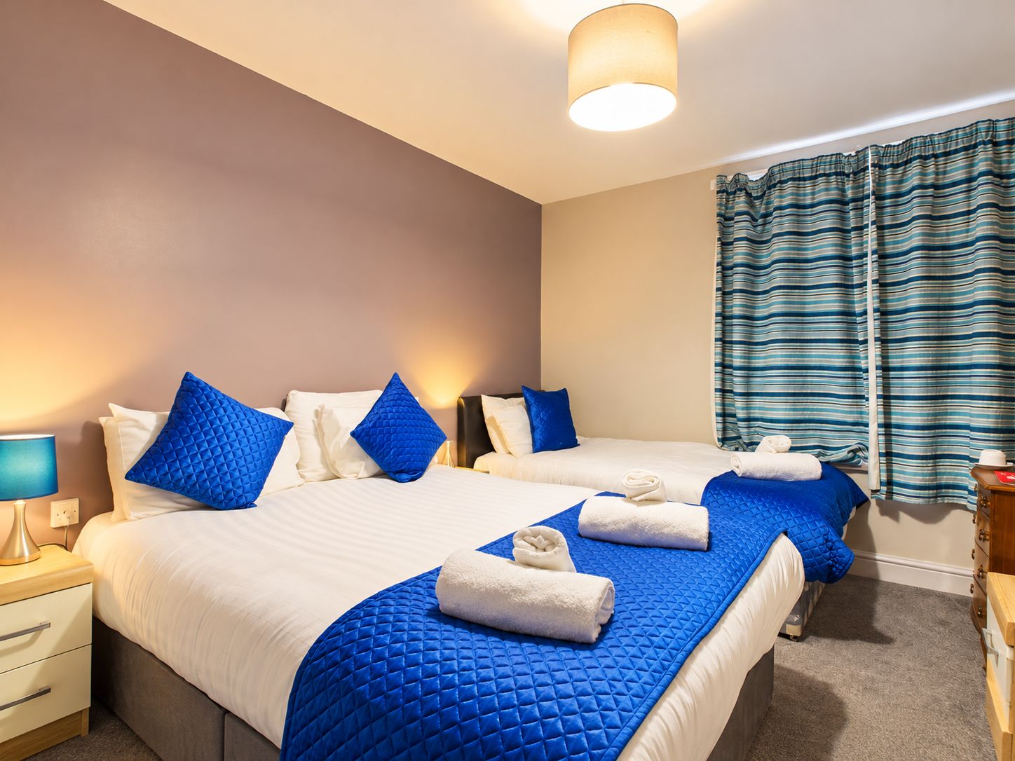 A hotel room with two beds, a nightstand with a lamp, a bedside table, and a window with curtains.