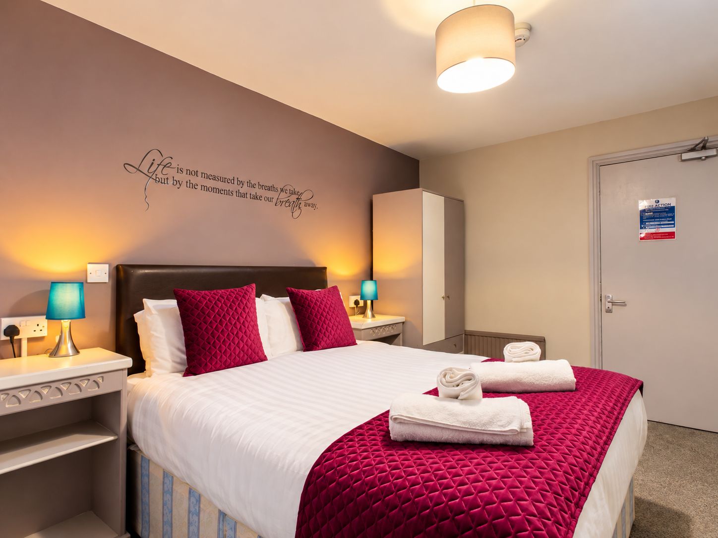 Habitación de hotel con una cama, dos lámparas de noche, un armario y una pared con una cita motivacional.