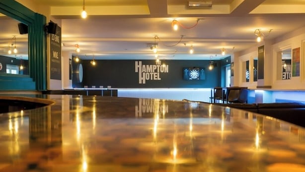 Un vestíbulo amplio con piso pulido que refleja las luces, un mostrador de recepción y un cartel de The Hampton Hotel.