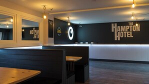 Un moderno vestíbulo de hotel con áreas de descanso, un mostrador de recepción y un gran cartel en la pared que dice "The Hampton Hotel".