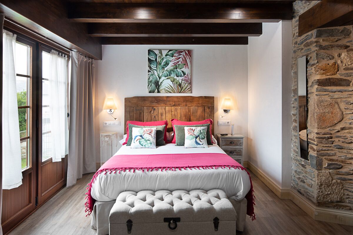 A bedroom with a wooden headboard, a large bed with pink and green pillows, a stone wall, and a window with curtains.