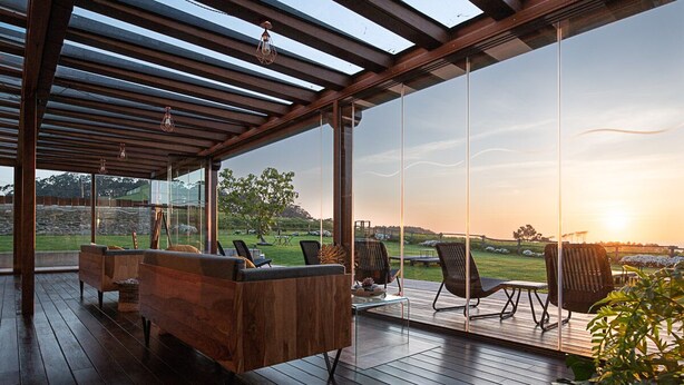Un espacio de estar al aire libre cubierto, con mobiliario de madera y vistas a un paisaje con césped al atardecer.