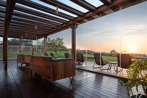 Un espacio de estar al aire libre cubierto, con mobiliario de madera y vistas a un paisaje con césped al atardecer.