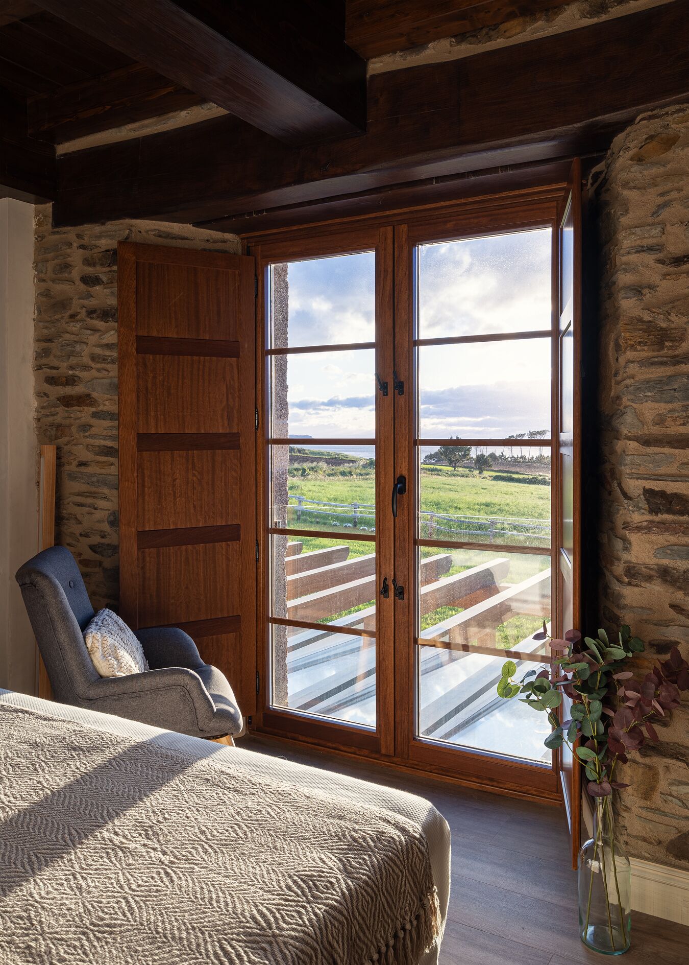 A bedroom with a large sliding glass door leading to a balcony, a wooden wardrobe, a cushioned chair, and a bed with a textured blanket.