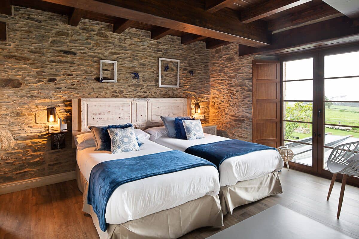 A bedroom with two beds, stone walls, wooden beams, and a large window with a view of greenery.