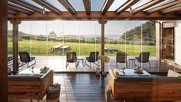 Una amplia terraza al aire libre con mobiliario de madera y paredes de vidrio que ofrece vistas a un paisaje verde y una masa de agua.