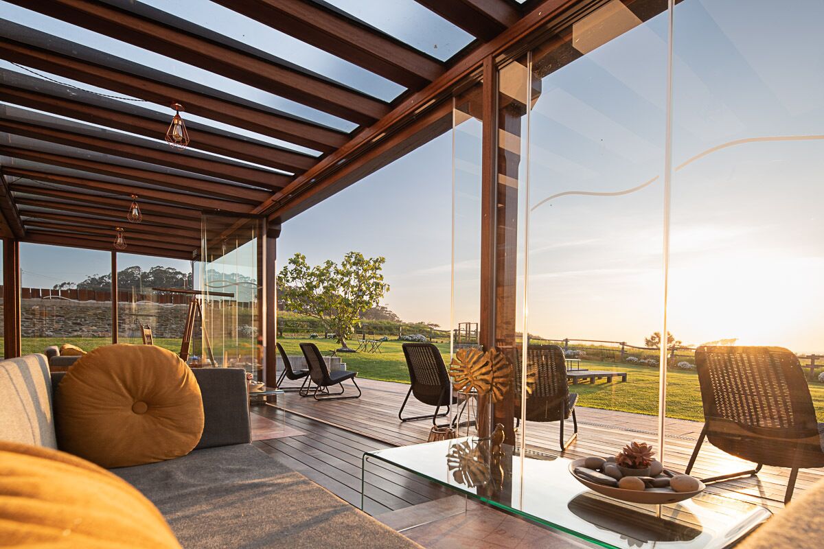 A modern outdoor seating area with glass walls, wooden flooring, and a view of a grassy landscape.