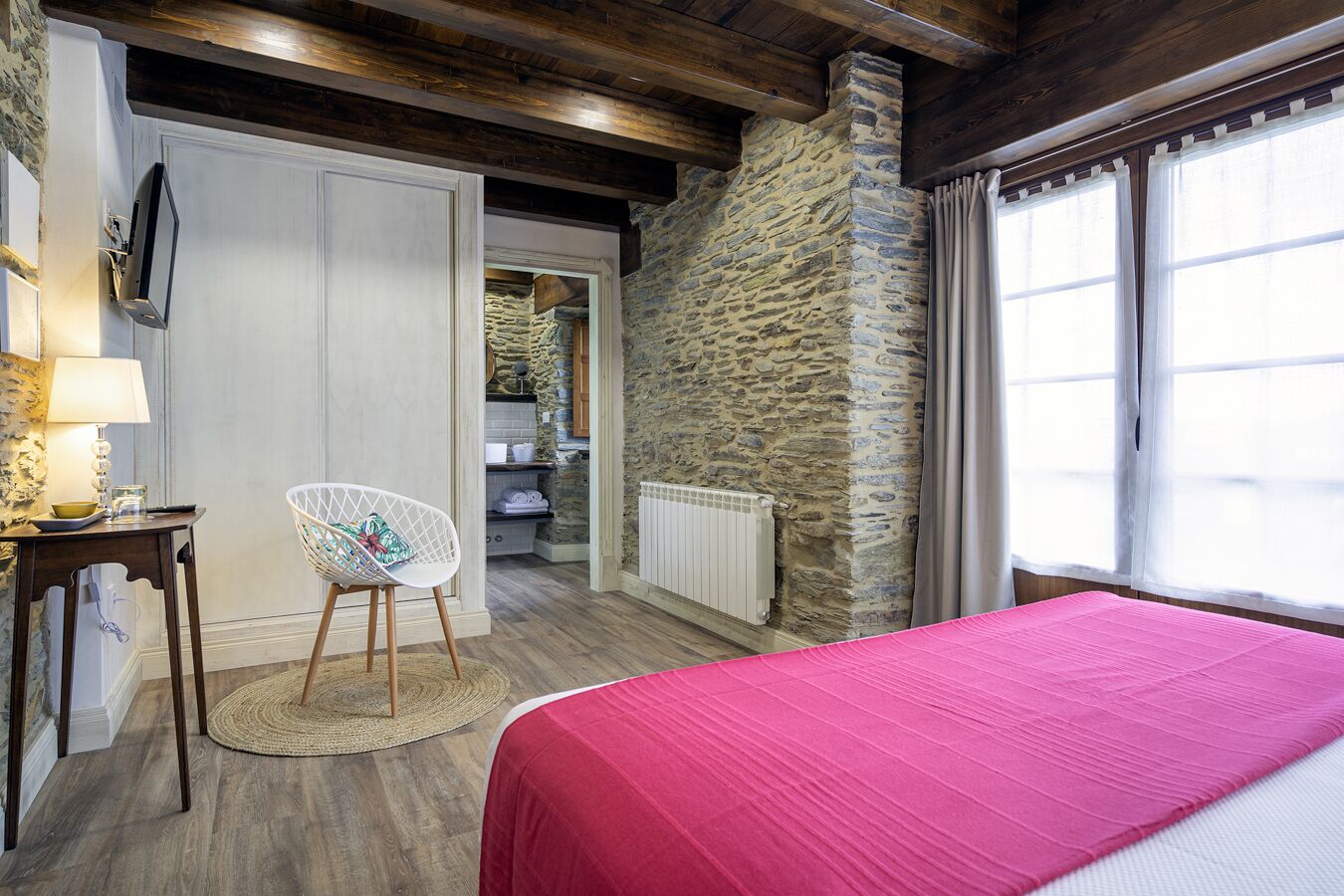 Un dormitorio con pared de piedra, cama con cubrecama rosa, techo de madera y una silla blanca.