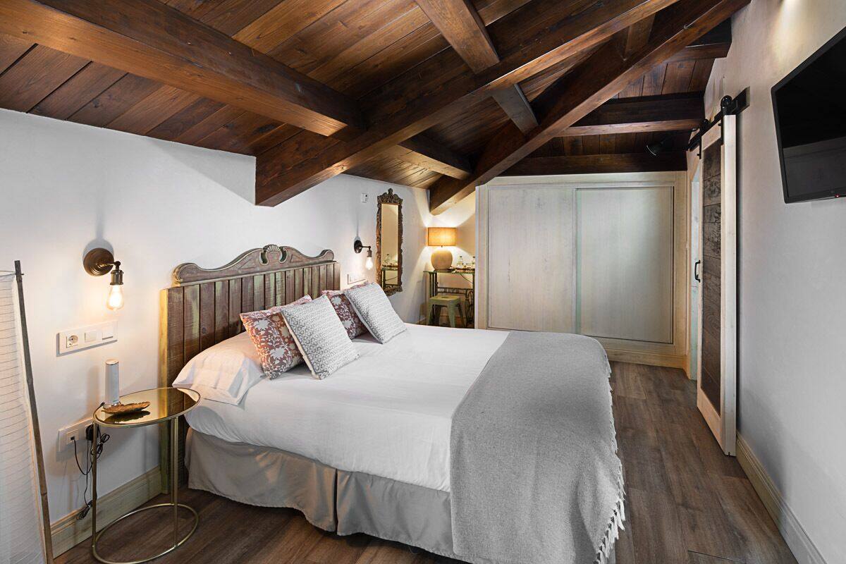 A bedroom with a wooden ceiling, a bed with multiple pillows, a bedside table, a lamp, and a wardrobe.