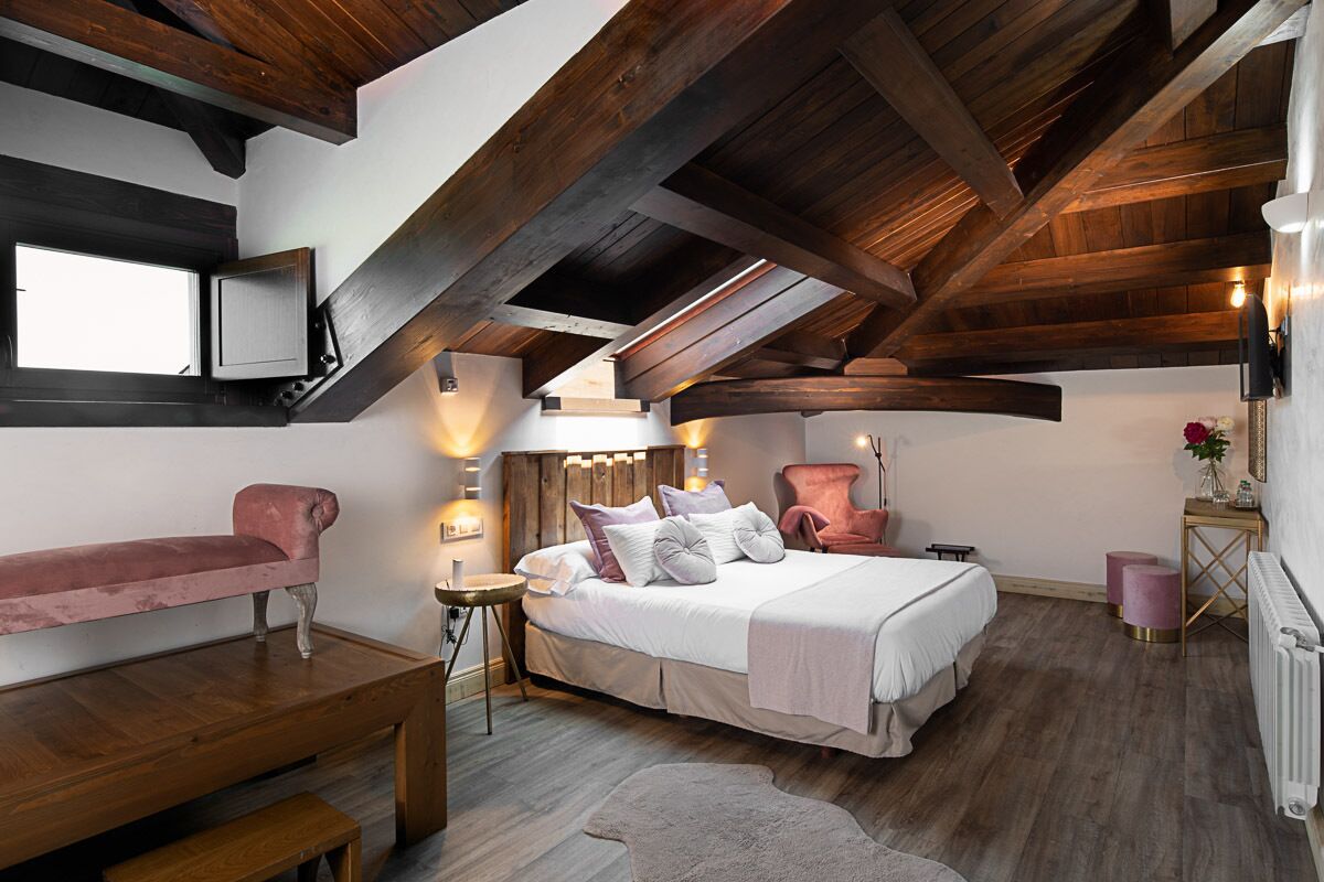 A bedroom with a wooden ceiling, a bed with pillows, a bench, a small table, and a radiator.