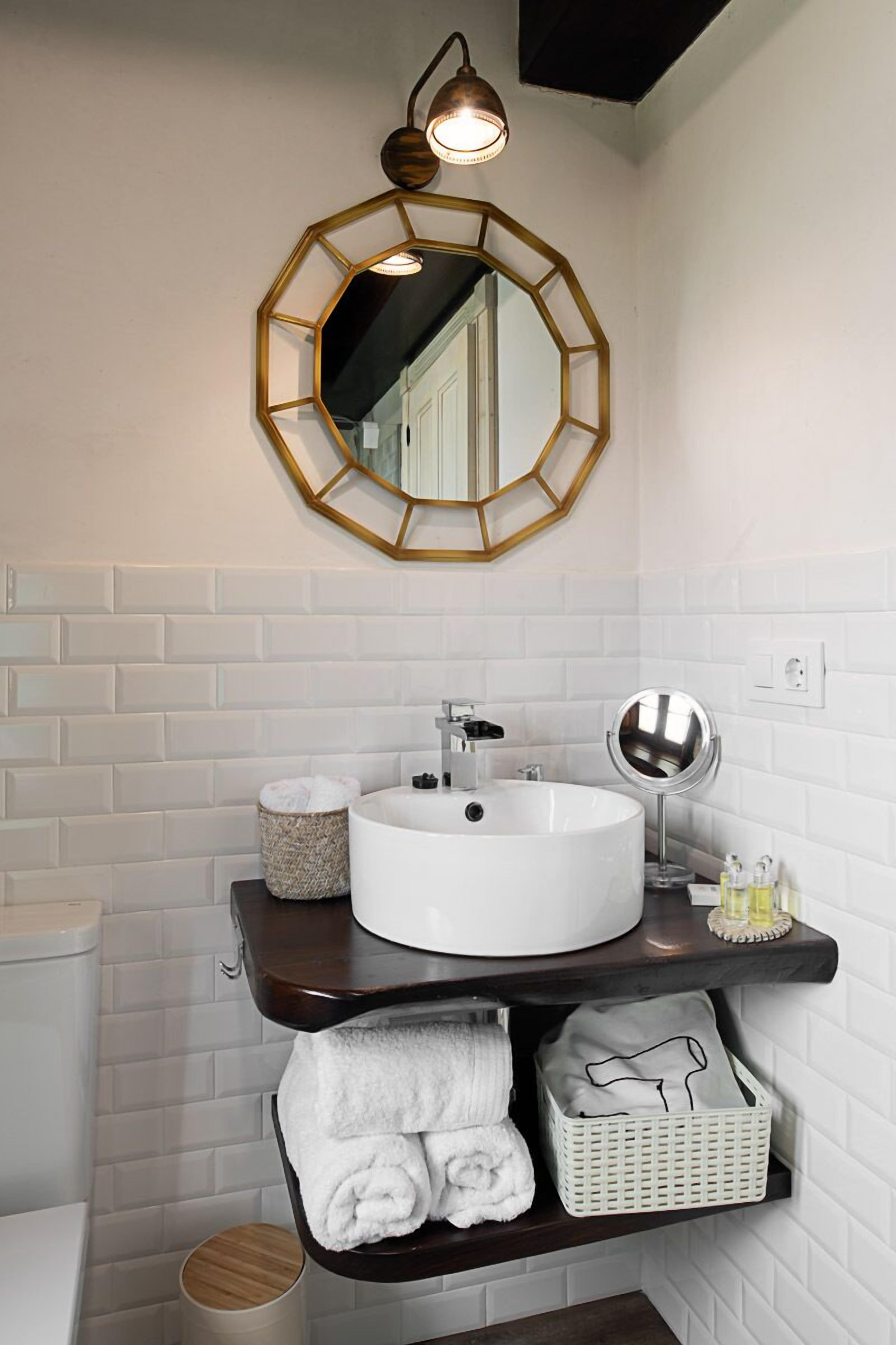 A bathroom with a white tiled wall, a white sink with a round mirror, and a wooden shelf with towels.