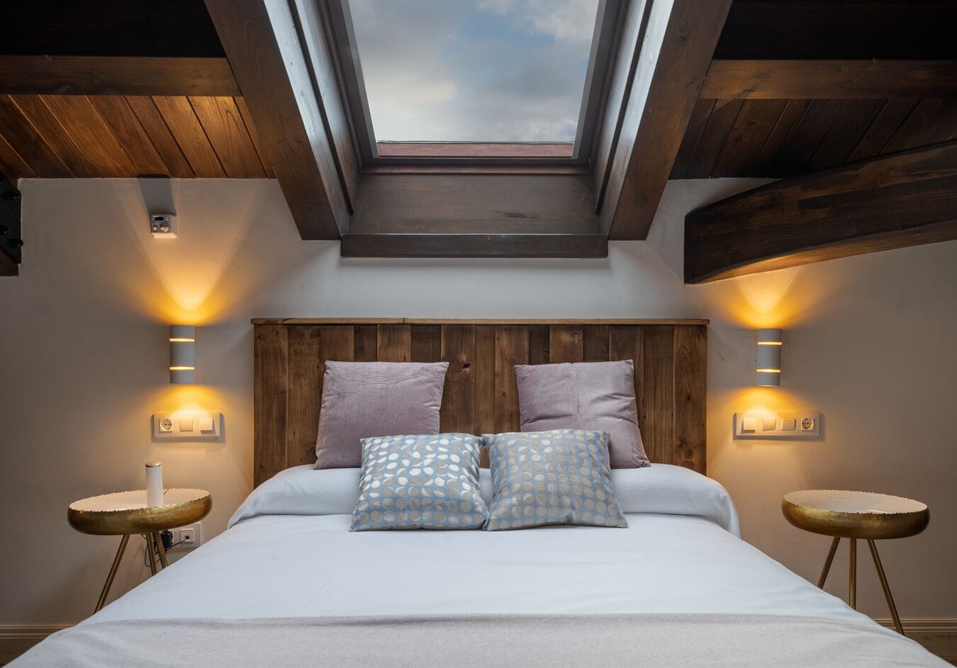 Un dormitorio con tragaluces, vigas de madera en el techo y una cama con dos almohadones, además de lámparas de noche.