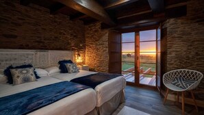 Un dormitorio con dos camas, una pared de piedra, techo de madera y vistas al atardecer a través de un ventanal.