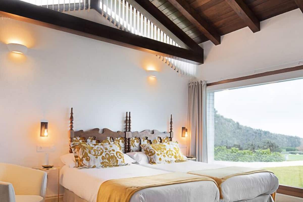 A bedroom with a wooden ceiling, a large bed with white bedding and yellow accents, a nightstand with a lamp, and a view of a green landscape through a large window.