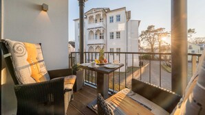 Un balcón con mobiliario de mimbre, una mesa con platos y frutas, y vista a un edificio blanco con balcones.