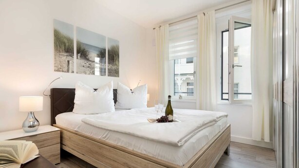 Un dormitorio moderno con una cama de madera, ropa de cama blanca, una mesita de noche con una lámpara y una obra de arte en la pared.