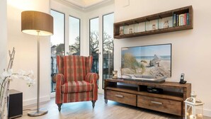 Una sala de estar con un sillón a cuadros, un televisor sobre un mueble de madera, una lámpara de pie y una estantería con libros y objetos decorativos.