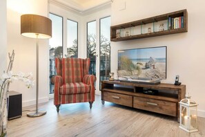 Un salon avec un fauteuil à carreaux, un téléviseur posé sur un meuble en bois, une lampe sur pied et une bibliothèque remplie de livres et d’objets décoratifs.