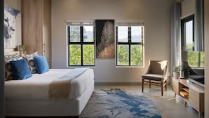 A modern bedroom with a large bed, a chair, and a view of greenery through the windows. - The Den Apartments (Stellenbosch)