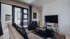 A modern living room with a sofa, a coffee table, a TV, and a large window with curtains. - The Den Apartments (Stellenbosch)