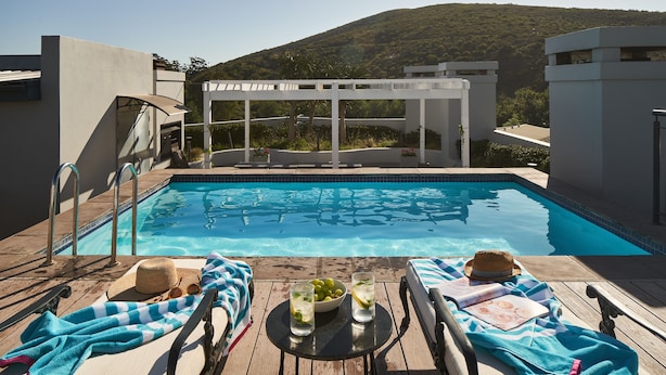 A swimming pool with lounge chairs and umbrellas, a table with drinks, and a view of a hillside. - The Den Apartments (Stellenbosch)