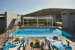 Piscina com espreguiçadeiras e guarda-sóis, uma mesa com bebidas e vista para uma encosta.