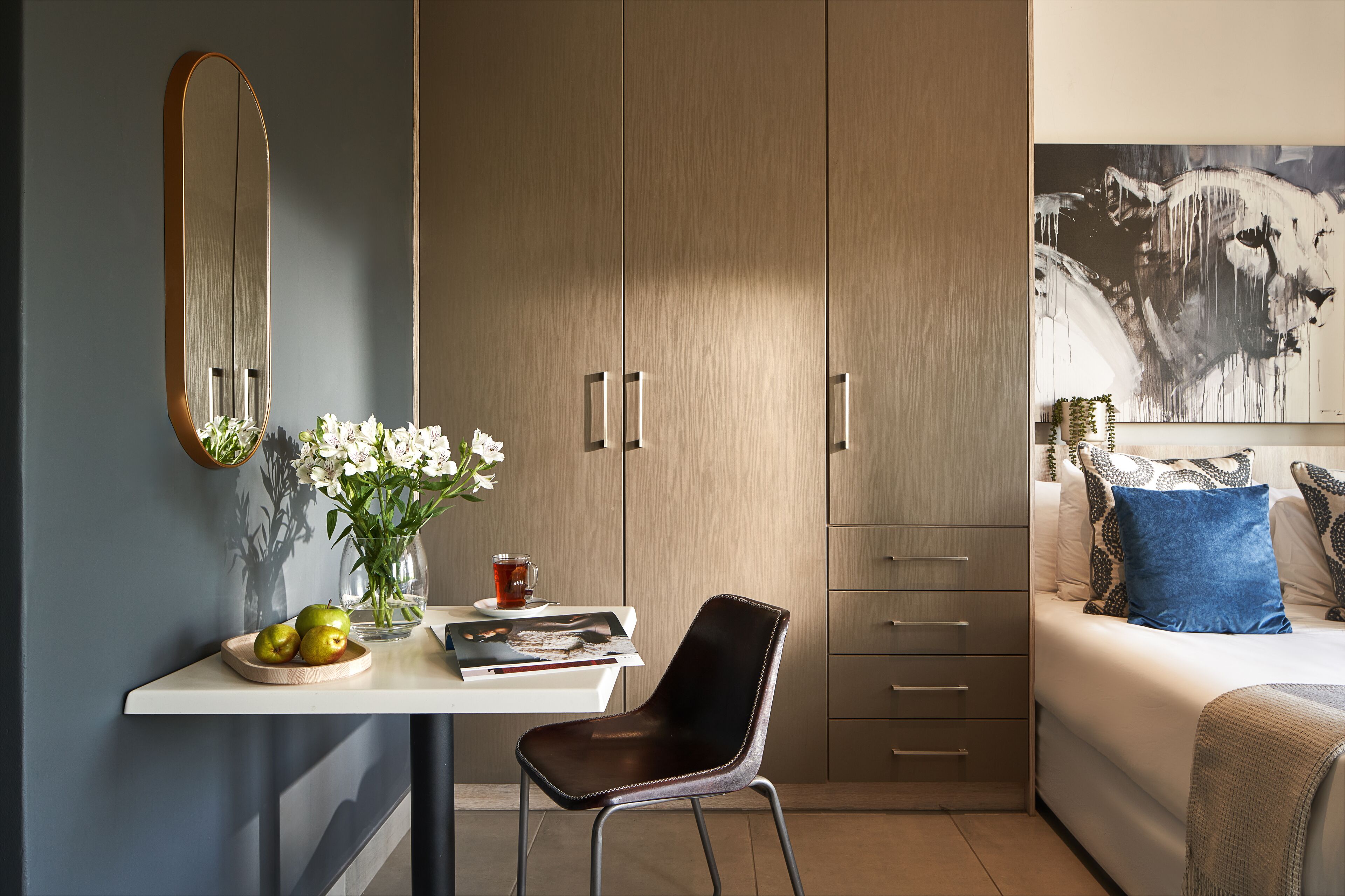 A modern bedroom with a bed, a desk with a chair, a wardrobe, and a large painting on the wall.