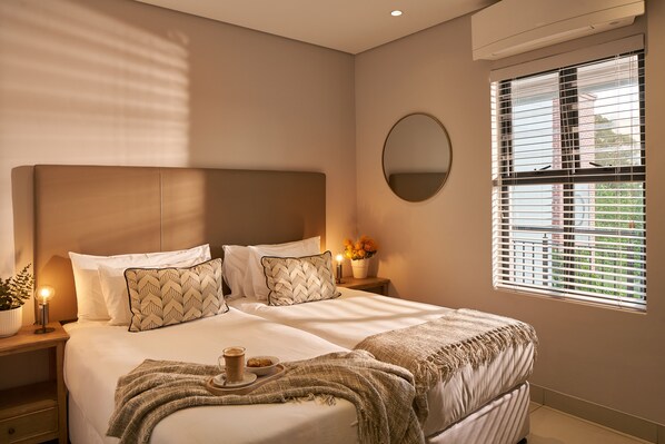A neatly made bed with pillows, a bedside table with a lamp, and a window with blinds.