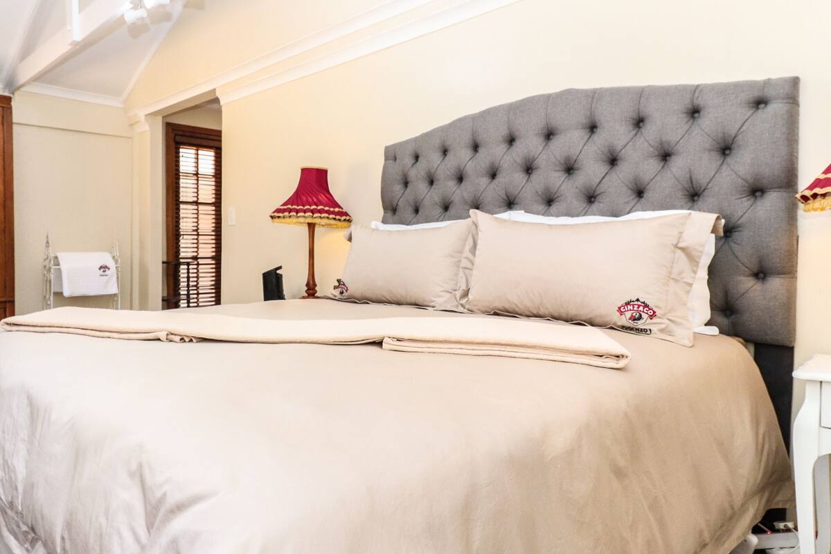 A neatly arranged bedroom with a large bed, a wooden wardrobe, a small table, and a lamp.