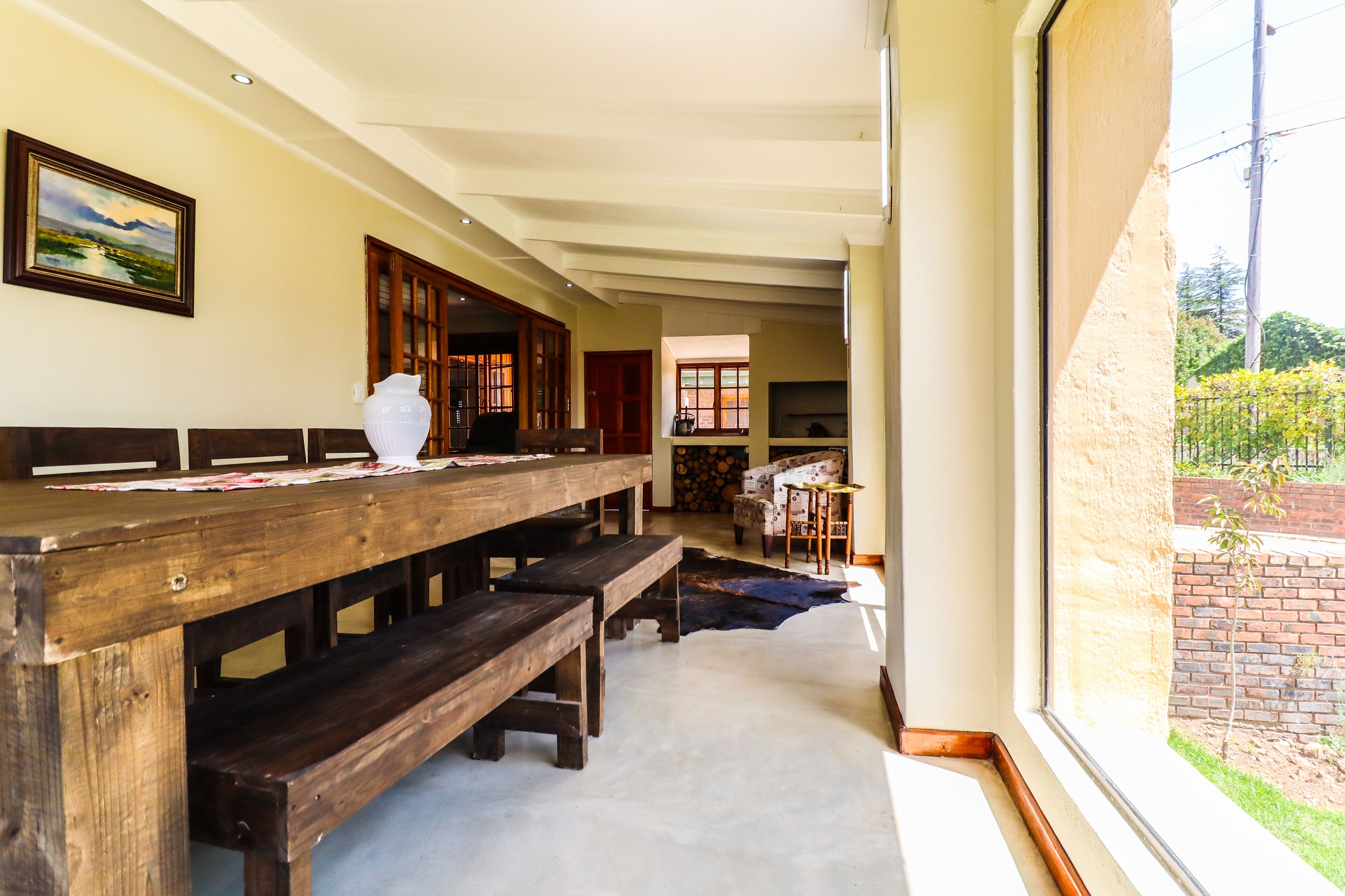 A dining area with wooden benches and a table, a framed painting on the wall, and a view of a brick wall and greenery outside.