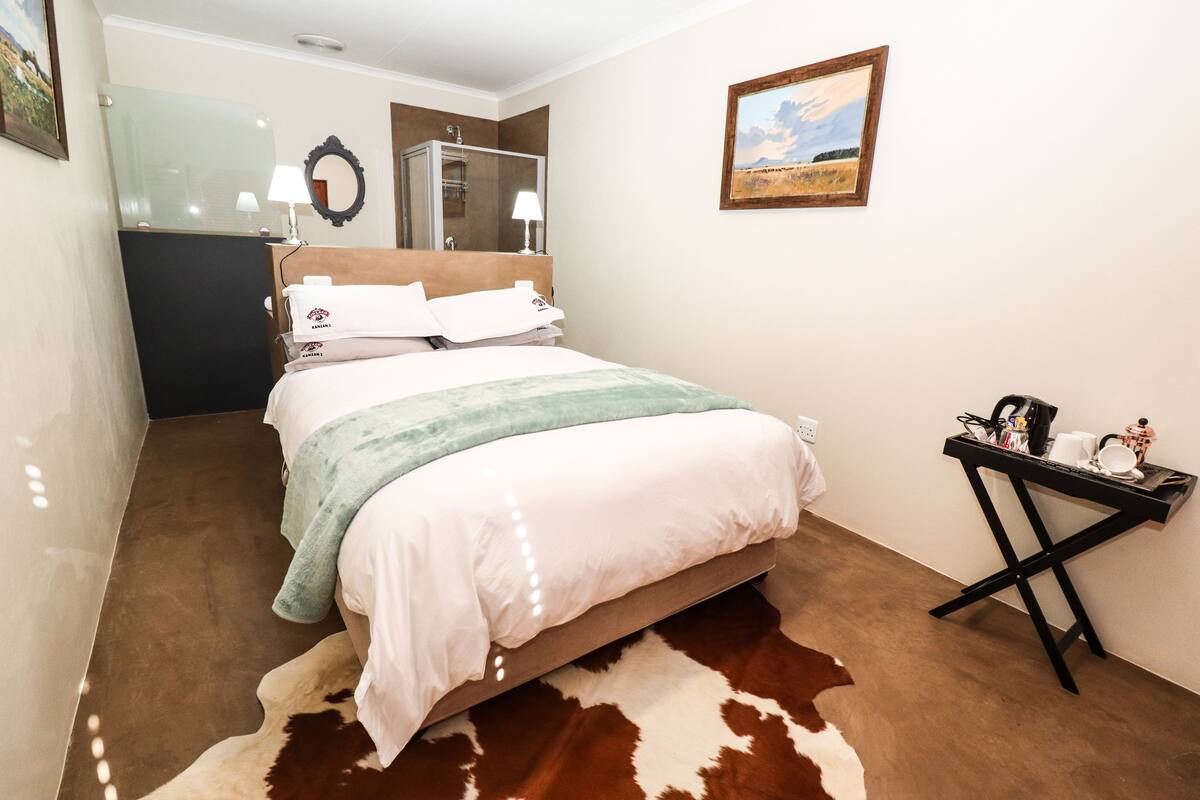 A hotel room with a bed, a small table with items, a mirror, and a painting on the wall.
