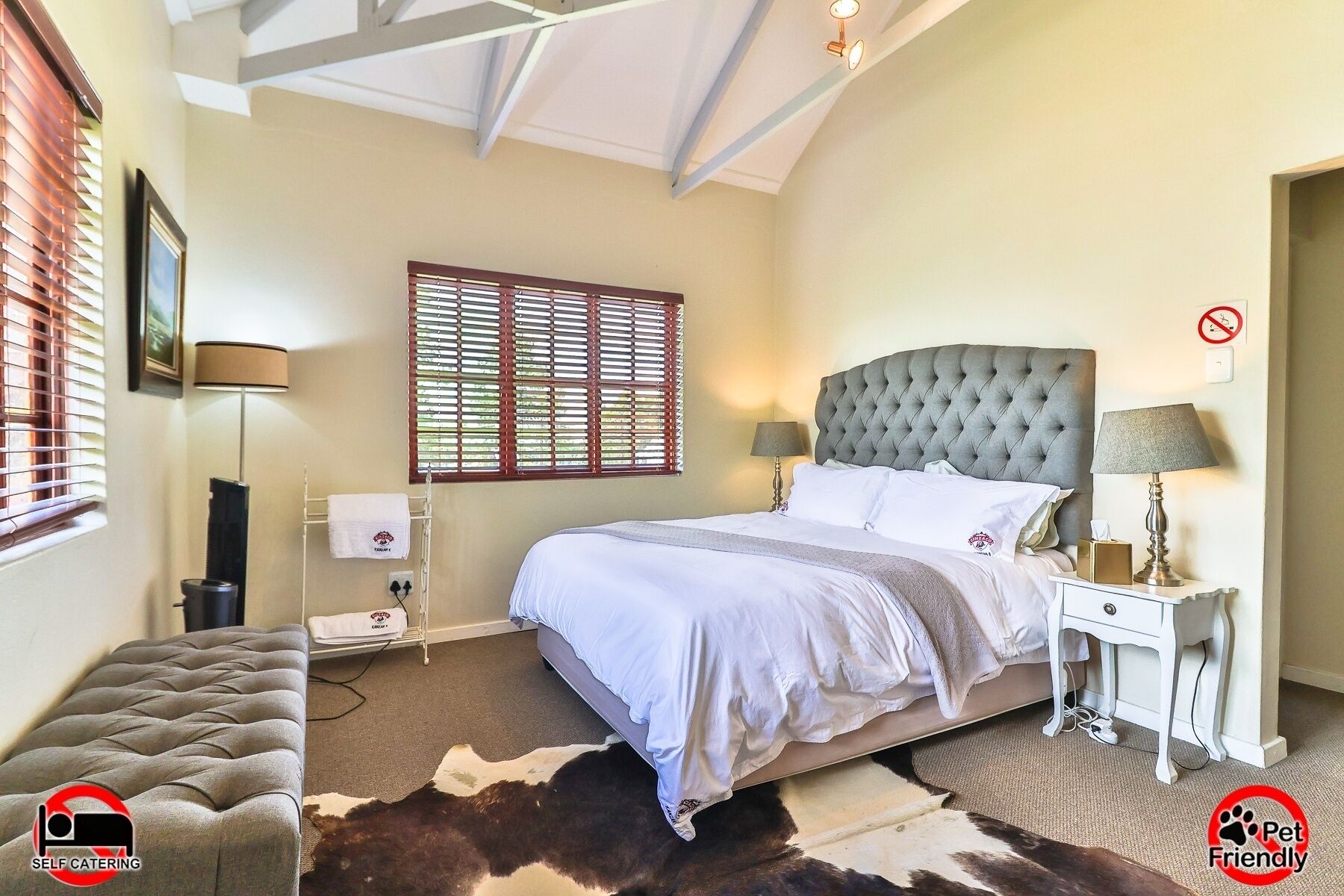 A bedroom with a large bed, a nightstand, a lamp, a window with blinds, and a cowhide rug.