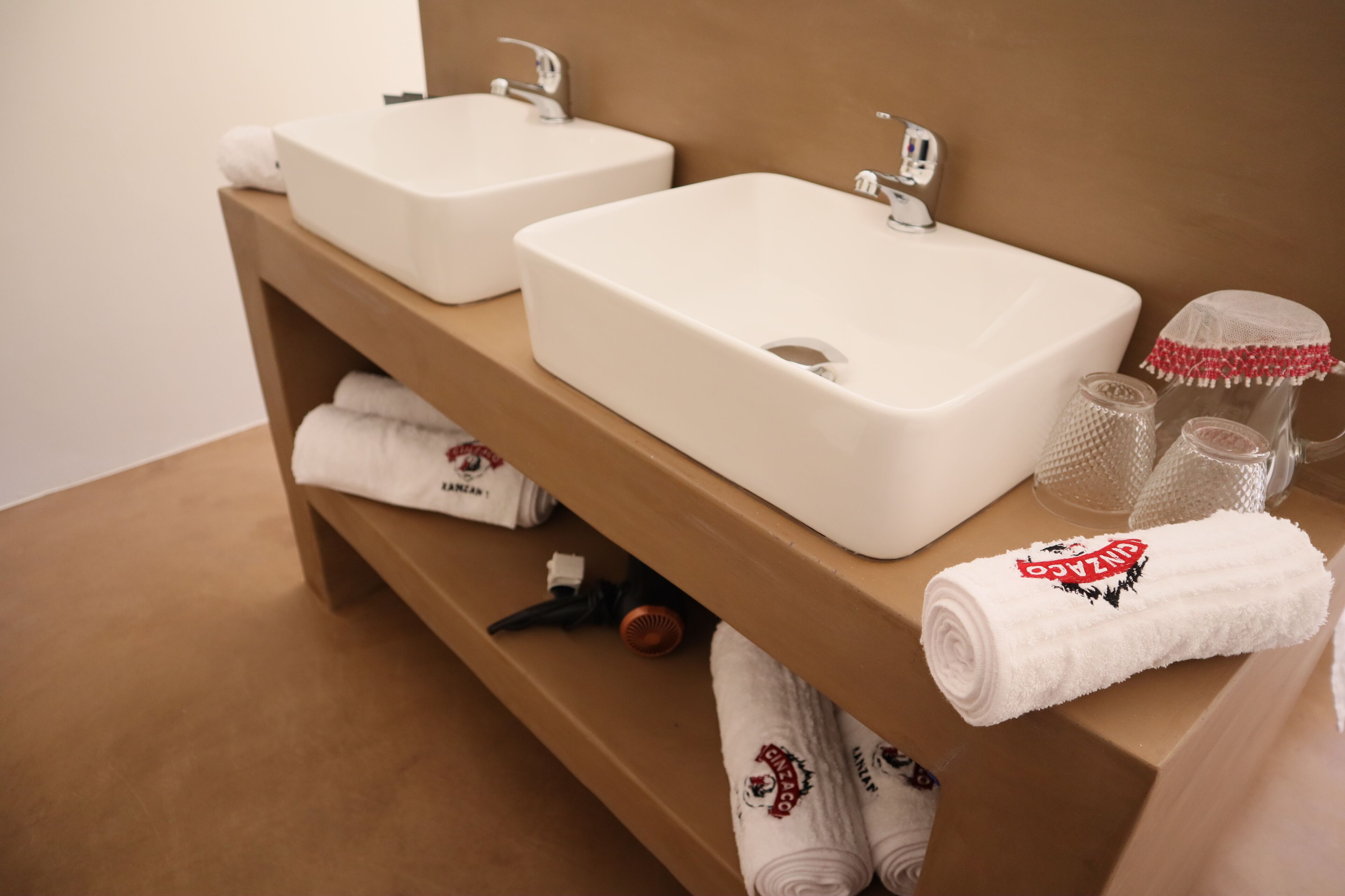 A bathroom with two white sinks, a wooden vanity, and neatly folded towels with a logo.