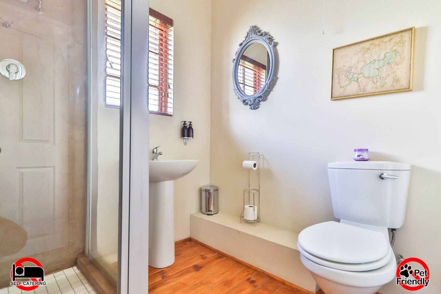 A bathroom with a toilet, sink, and mirror. There is a map on the wall and a window with blinds.