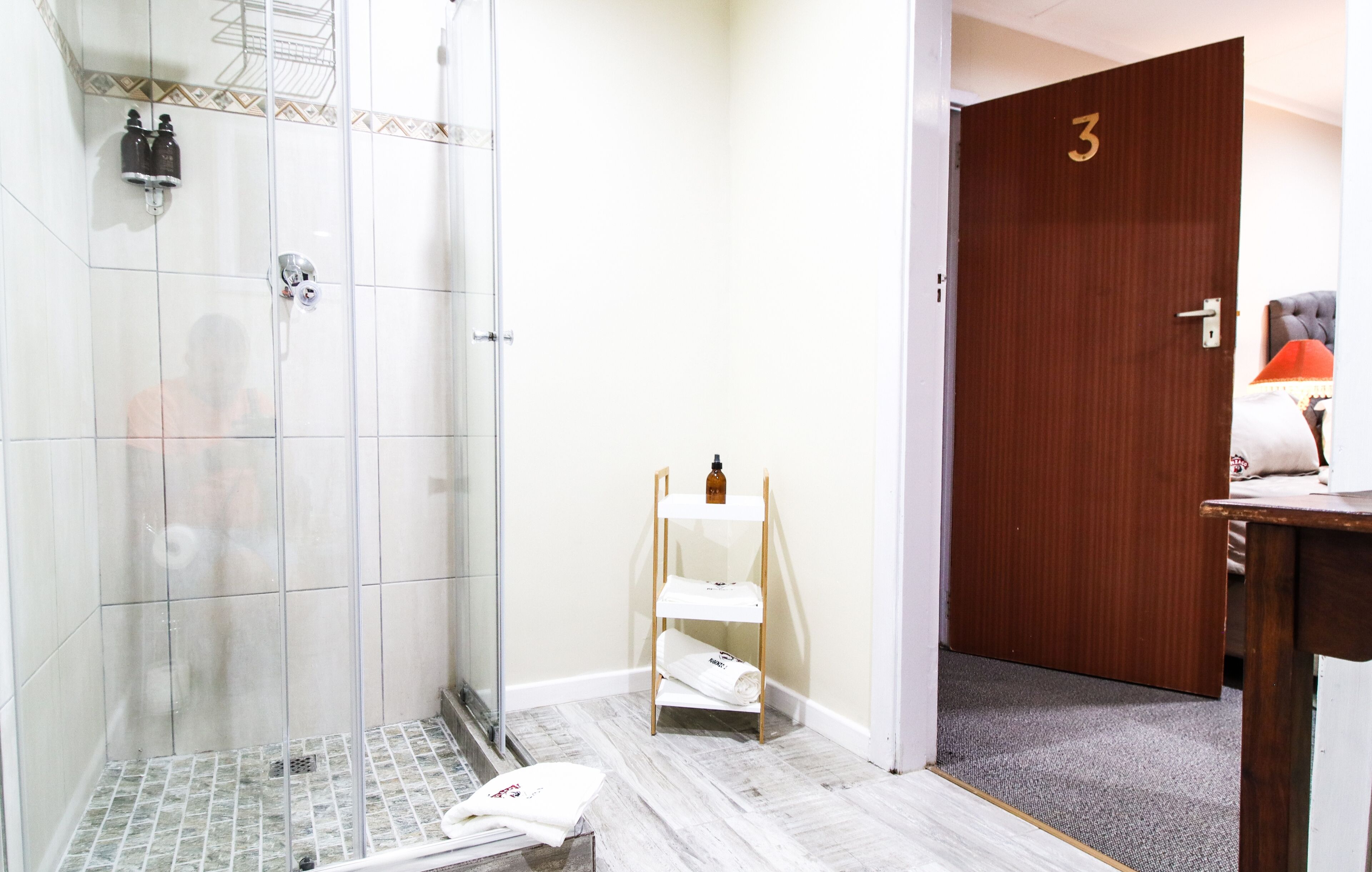 A hotel bathroom with a glass shower, a wooden door marked with the number 3, and a small shelf with towels.