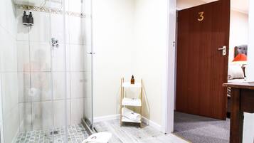 A hotel bathroom with a glass shower, a wooden door marked with the number 3, and a small shelf with towels.